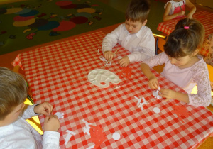Trójka dzieci przygotowuje kotylion z pasków papieru, sklejają elementy.